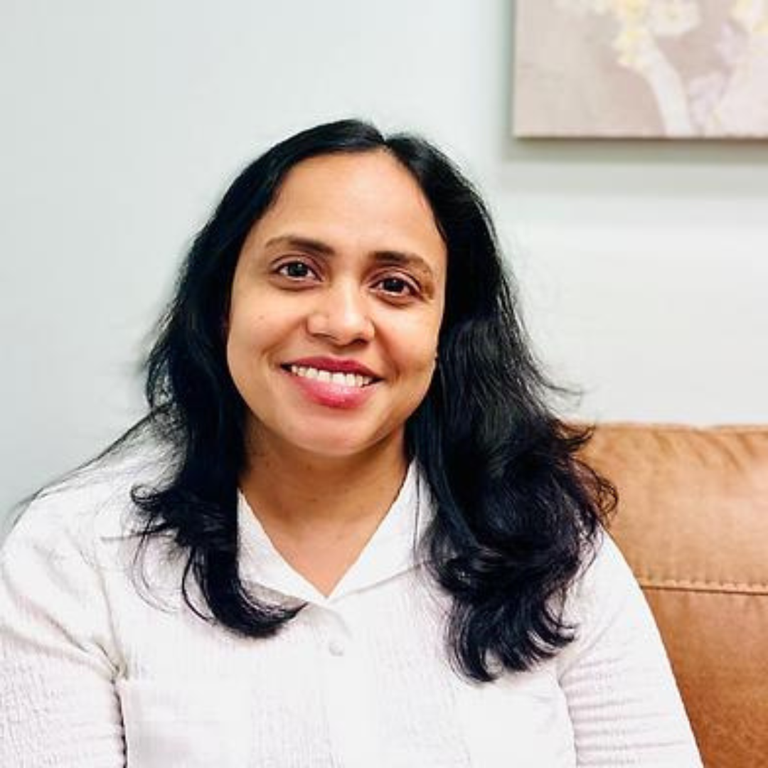 Smiling female therapist with long dark hair seated in a cozy indoor setting.