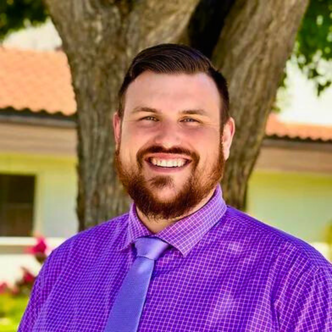 Smiling male therapist with a beard and wearing a purple shirt and tie, standing outdoors near a tree.