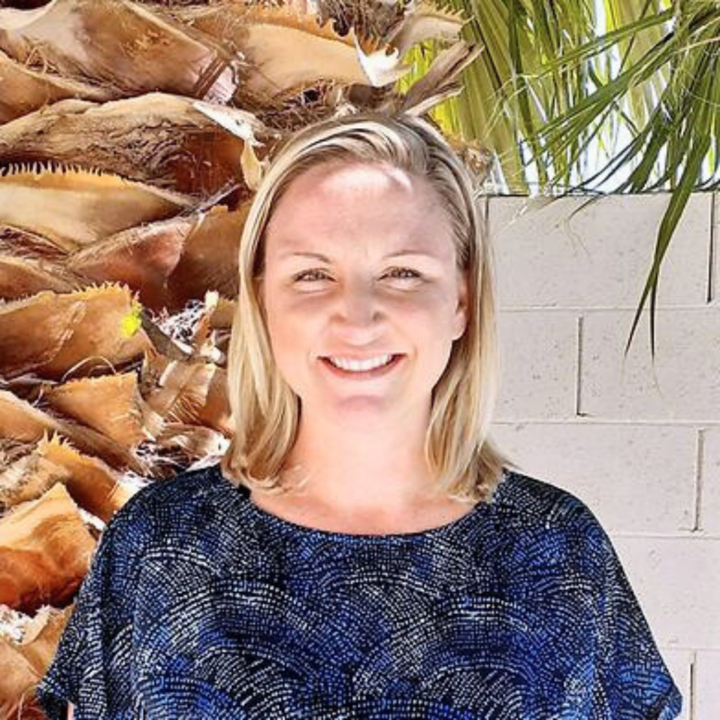 Smiling female therapist with blonde hair standing outdoors in front of a palm tree.