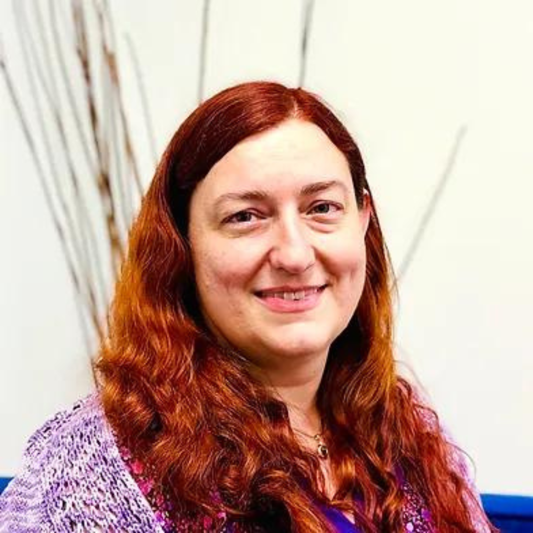 Smiling female therapist with long red hair seated indoors, wearing a purple sweater.