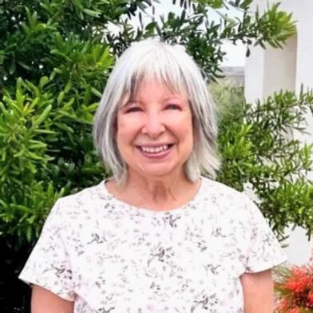 Smiling female therapist with short gray hair standing outdoors in front of greenery.