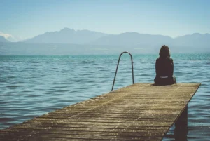 A person sits at the edge of a wooden dock overlooking a serene lake surrounded by mountains, appearing deep in thought.