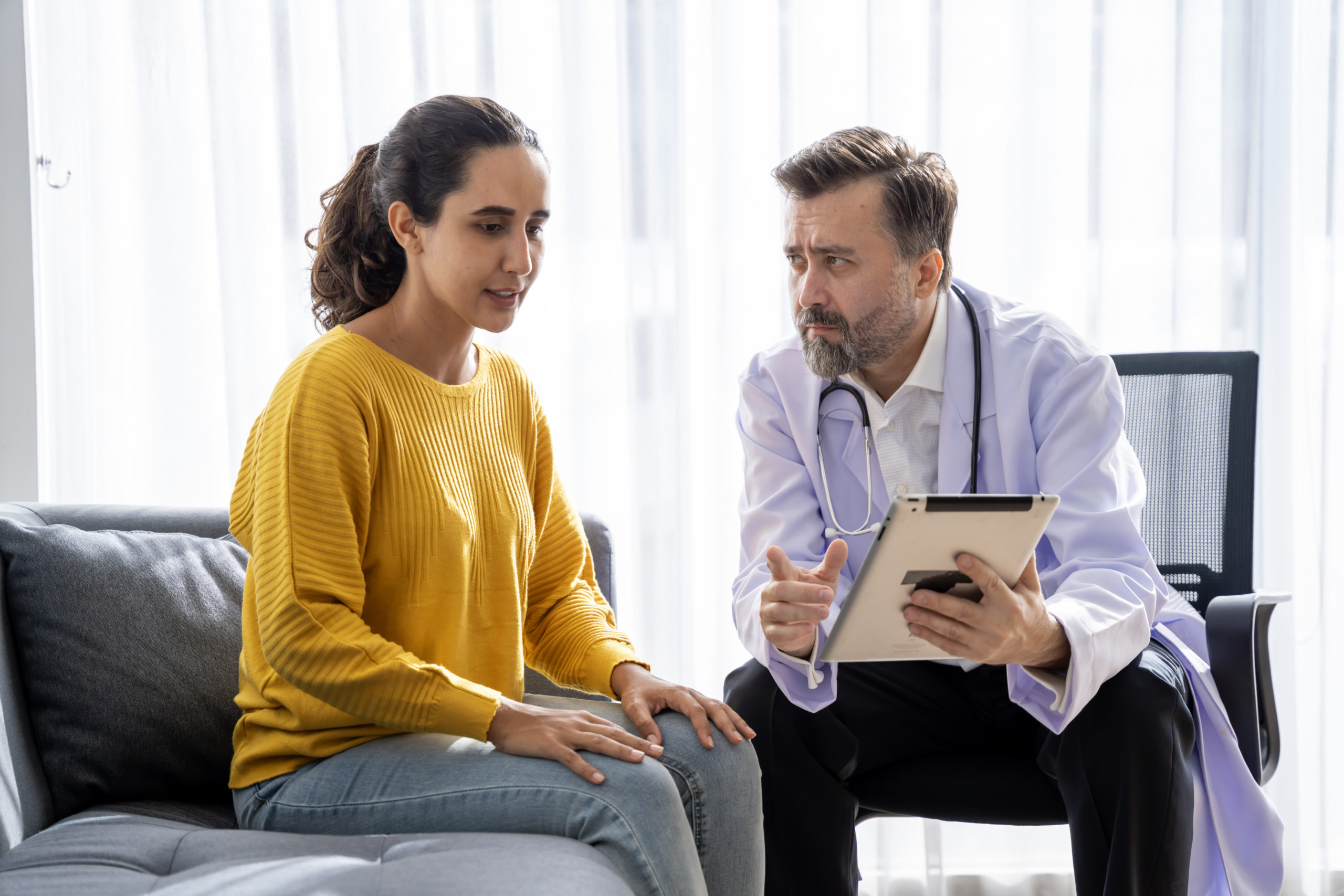 Male doctor attentively listening to a female patient while holding a tablet during a consultation.