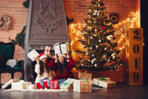 Two girls with cheerful expressions, presenting wrapped gifts in front of a sparkling Christmas tree