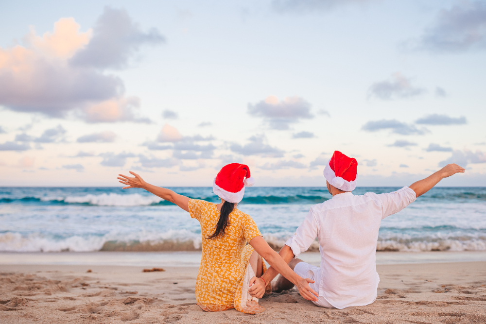 A couple wearing Santa hats enjoys a sunny day sitting together on the beach, surrounded by sand and ocean waves