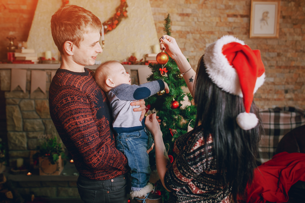 A couple stands with their baby, surrounded by a festive Christmas tree, capturing a moment of love and celebration