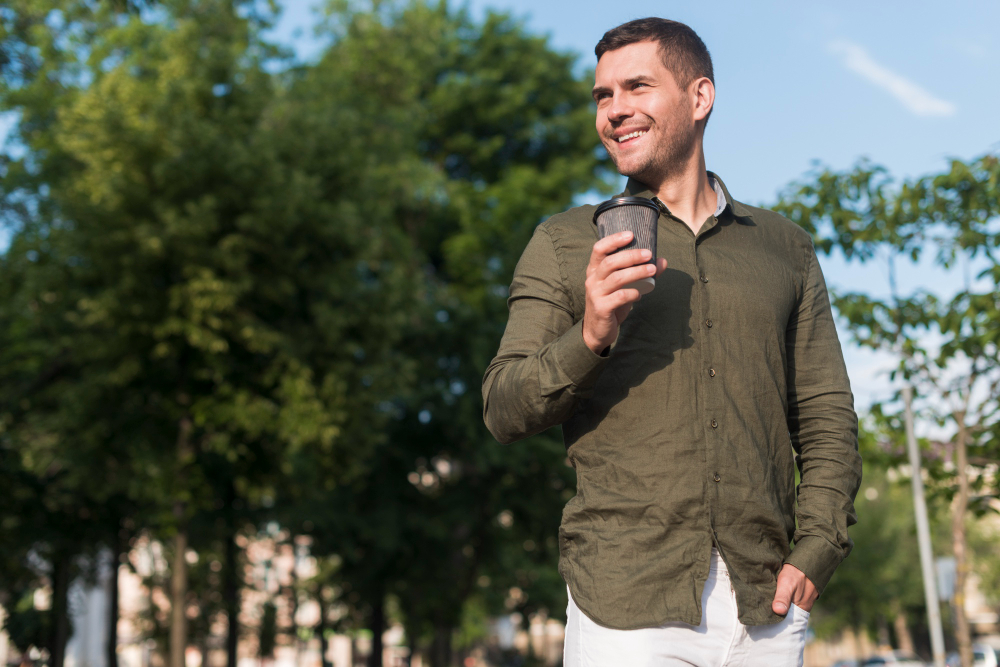 A man in a green shirt and white pants holds a cell phone, focused on the screen while standing outdoors