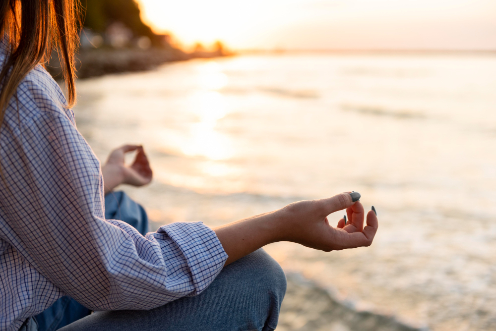 A woman sits cross-legged on the beach, meditating peacefully as the sun sets, casting warm hues across the sky.