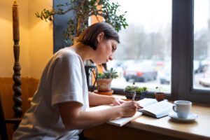 A woman seated at a table, writing in a notebook while enjoying a cup of coffee