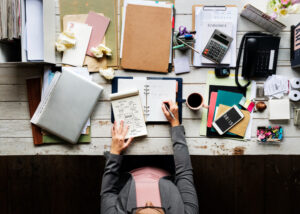 A busy professional seated at a desk, stressed and surrounded by papers and various items, focused on her work in a cluttered workspace