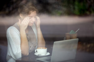 A sad woman seated at a table, working on a laptop while enjoying a cup of coffee