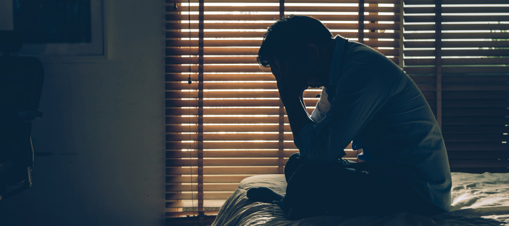 A man sits on a bed, his head resting in his hands, conveying a sense of distress and contemplation.