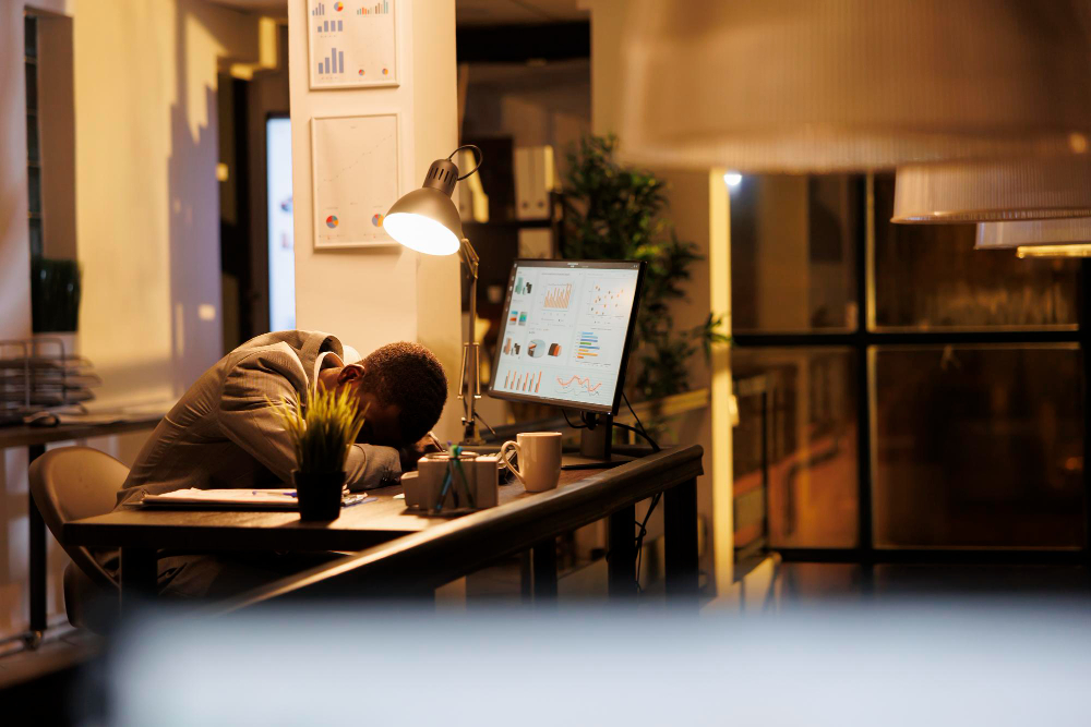 A burned-out man is slumped over a desk, asleep, with the soft glow of night illuminating the scene.