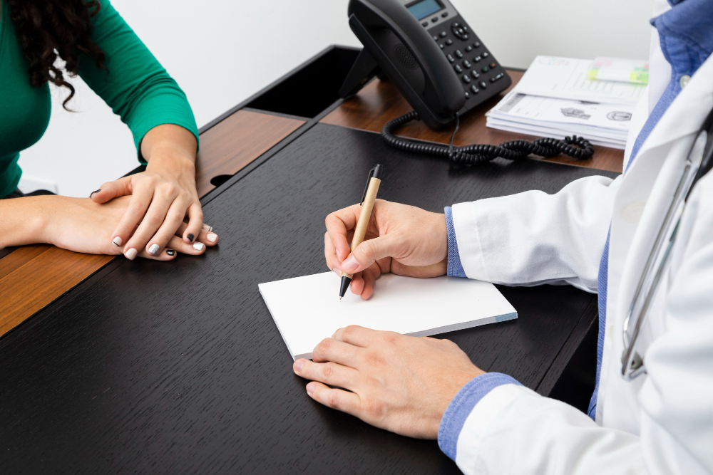 A doctor writes notes on a notepad while a patient sits at a desk, engaged in a consultation.