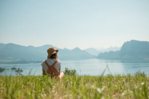 A woman with depression sits on a hill, gazing at a serene lake surrounded by majestic mountains in the background Seeking depression treatment at Lighthouse Psychiatry