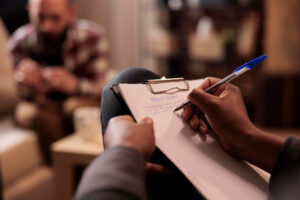 A person seated on a couch writes on a clipboard, undergoing Psychotherapy and Medication in a cozy indoor setting.