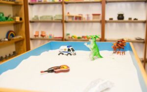 A toy car and a toy dinosaur are placed together in a sandbox, surrounded by fine grains of sand.