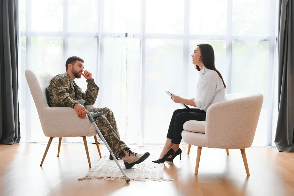 Veteran in military uniform using a crutch during a psychotherapy session with a female therapist discussing PTSD trauma recovery and mental health support in a bright counseling office